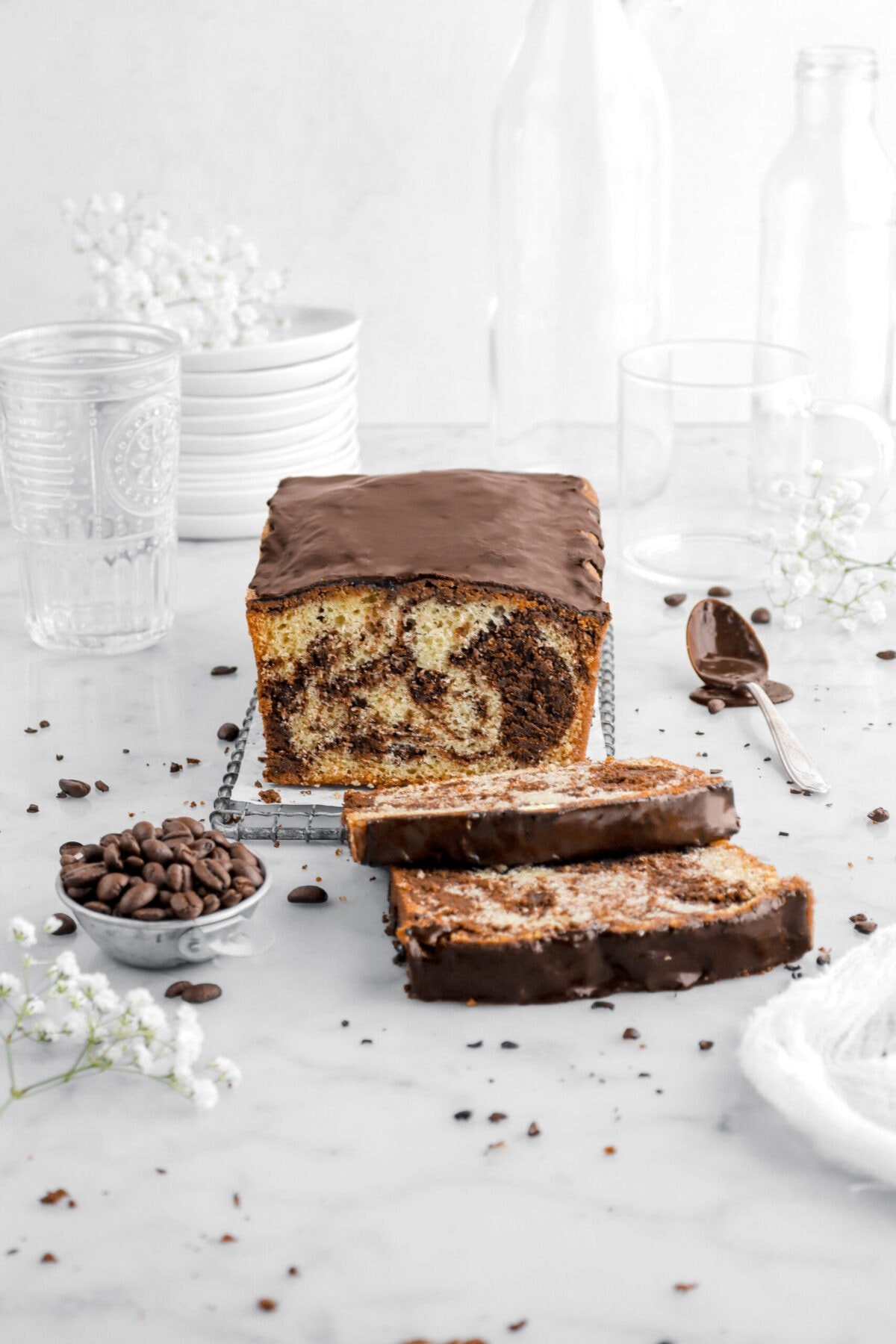 loaf cake with two slicing laying in front with chocolate icing on top with coffee beans and white flowers around, empty glasses and stack of plates behind.