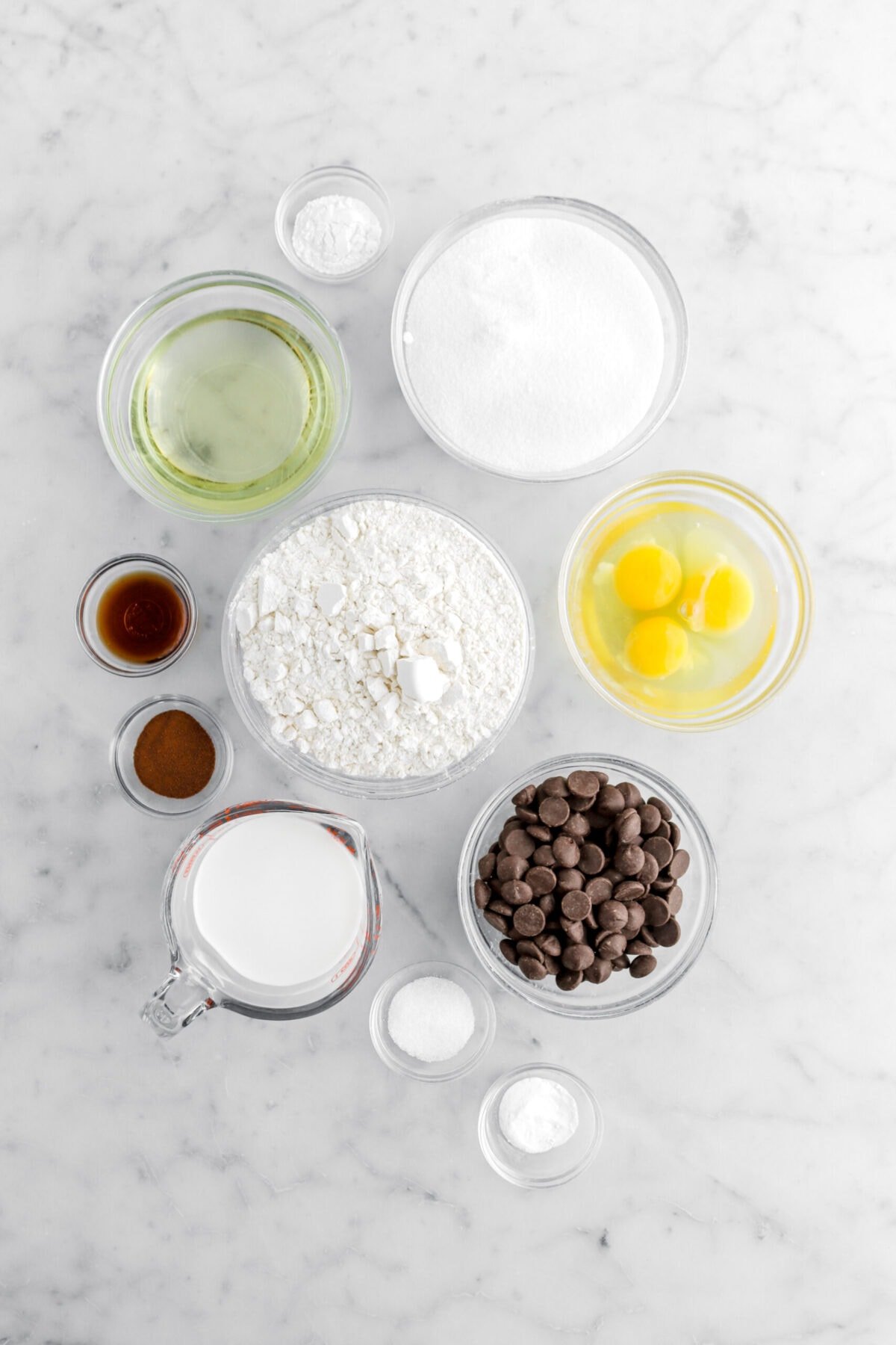 baking powder, sugar, vegetable oil, flour, vanilla, espresso powder, eggs, milk, chocolate chips, salt, and baking soda on marble surface.