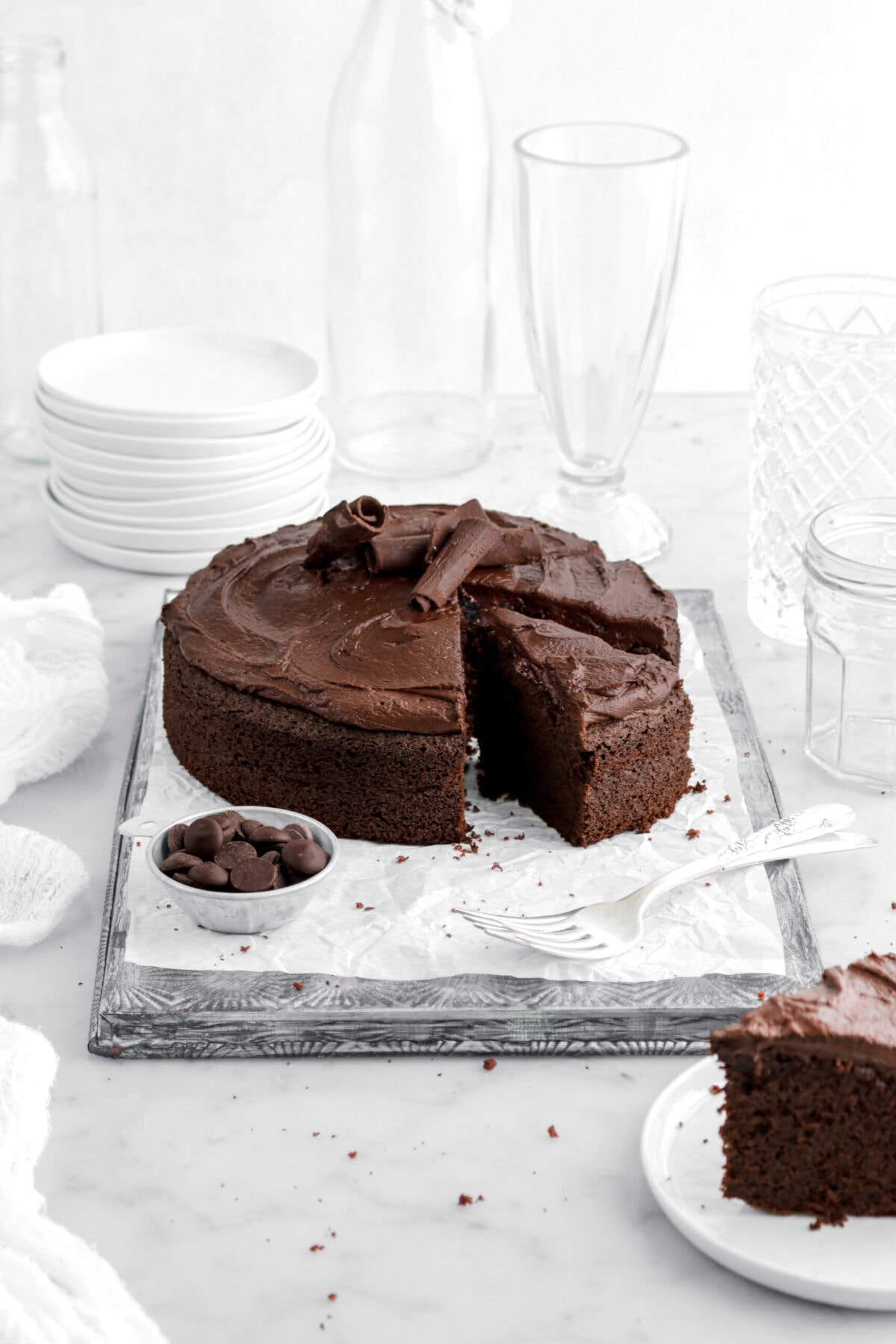 front shot of sliced chocolate fudge cake on upside down sheet pan with bowl of chocolate chips and forks beside and a plate with a slice of cake on white plate in front of cake with stack of plates and empty glasses behind on marble surface.