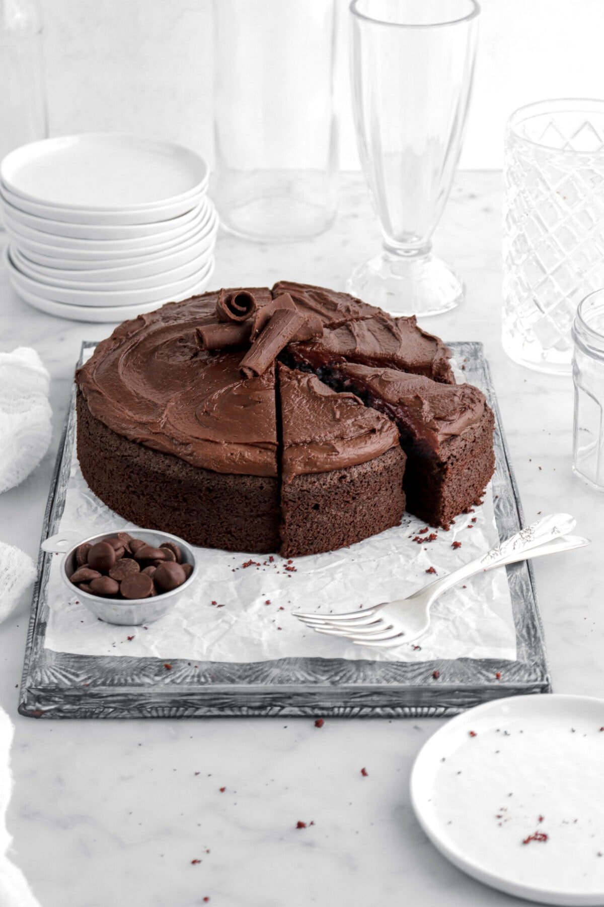 chocolate fudge cake with four slices cut into it with frosting and chocolate curls on top with bowl of chocolate chips and a fork in front of cake on lined sheet pan with plate in front and empty glasses behind.