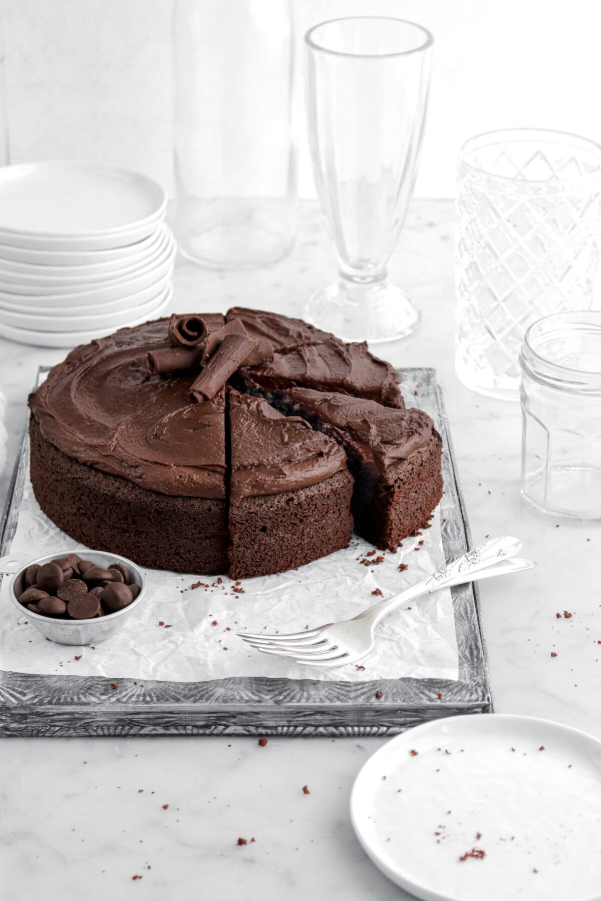 cropped image of chocolate fudge cake with four slices cut into it with bowl of chocolate chips and two forks in front.