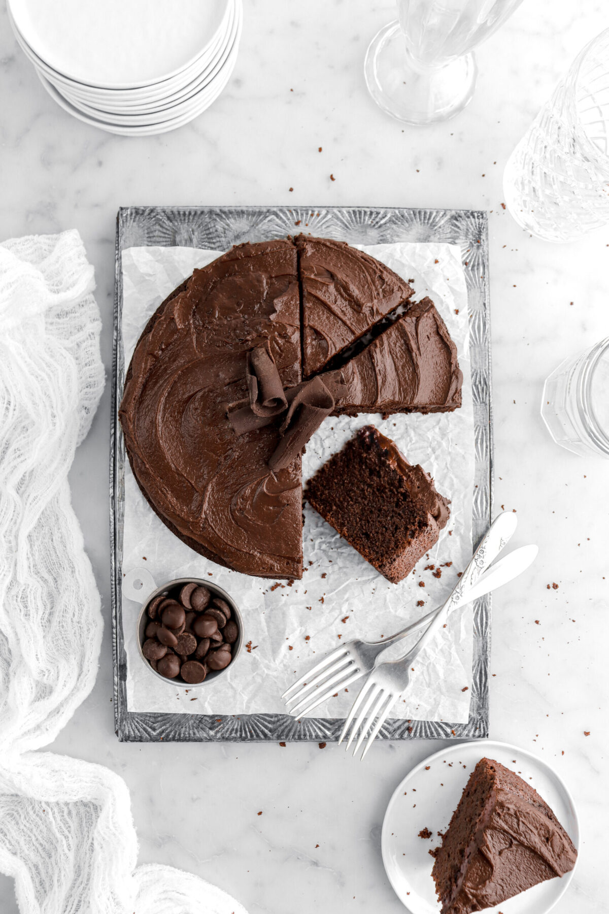 overhead image of chocolate cake with three slices next to cake with one turned on its side, a measuring cup of chocolate chips, two forks, and a slice of cake on white plate on marble surface.