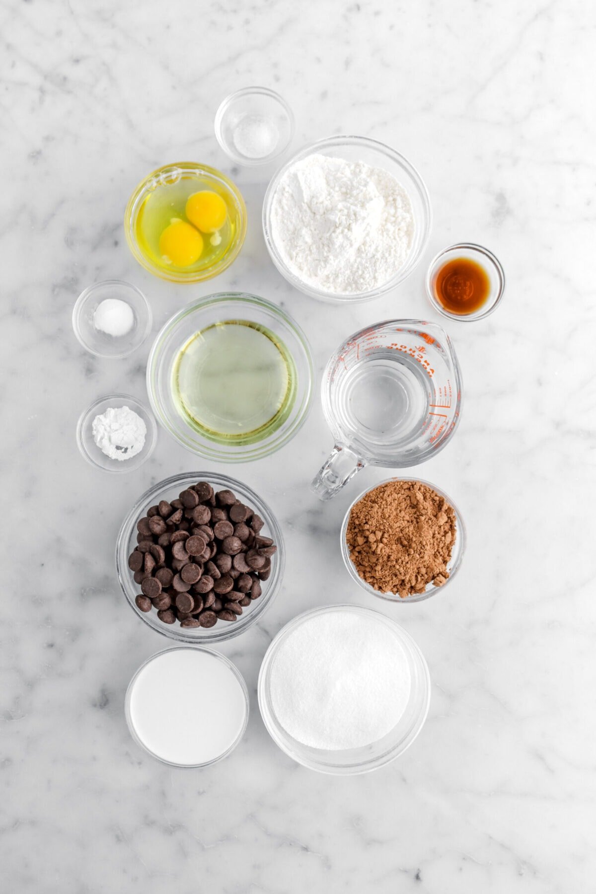 salt, eggs, flour, vanilla, baking soda, baking powder, oil, water, cocoa powder, chocolate chips, milk, and sugar on marble surface.