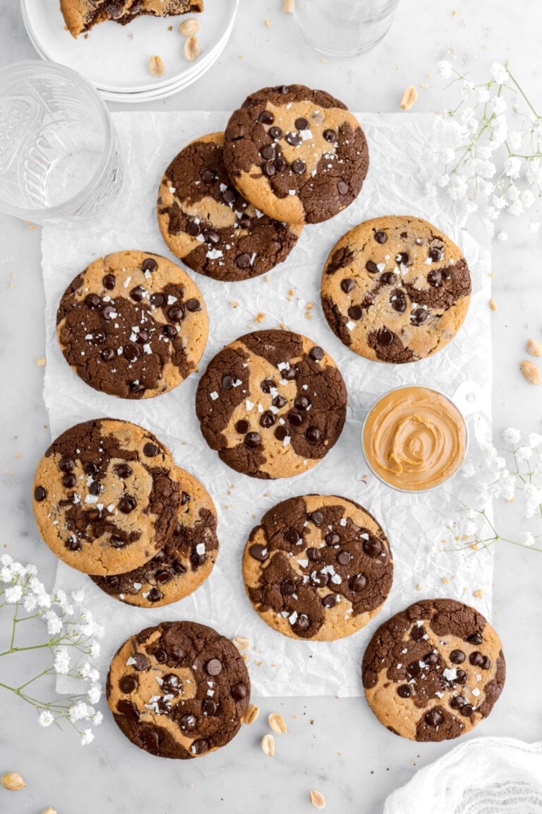 ten peanut butter chocolate marble cookies on parchment paper with flaked salt on top of each cookie with measurring cup of peanut butter beside and white flowers around.