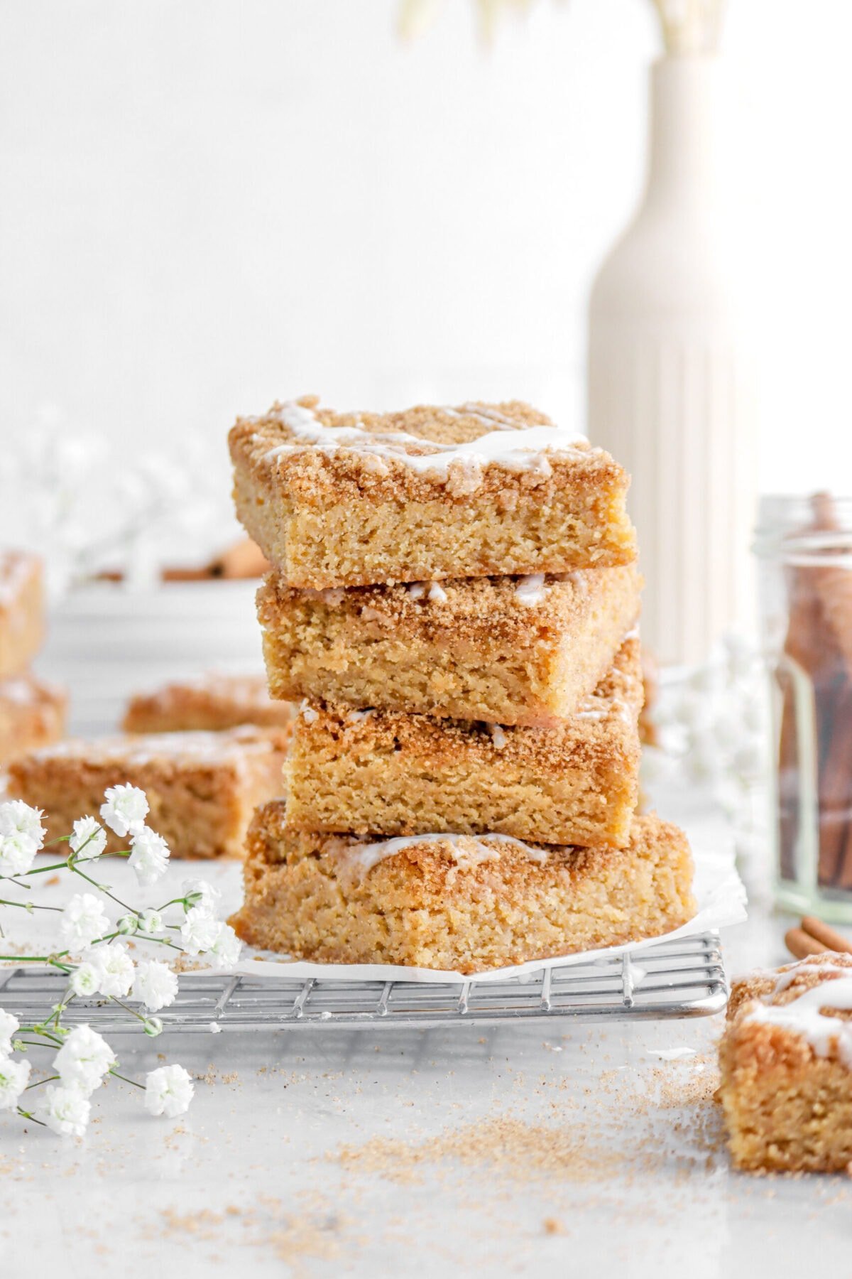 four stacked cinnamon roll blondies in wire cooling rack with white flowers beside, more blondies around, and a vase behind.