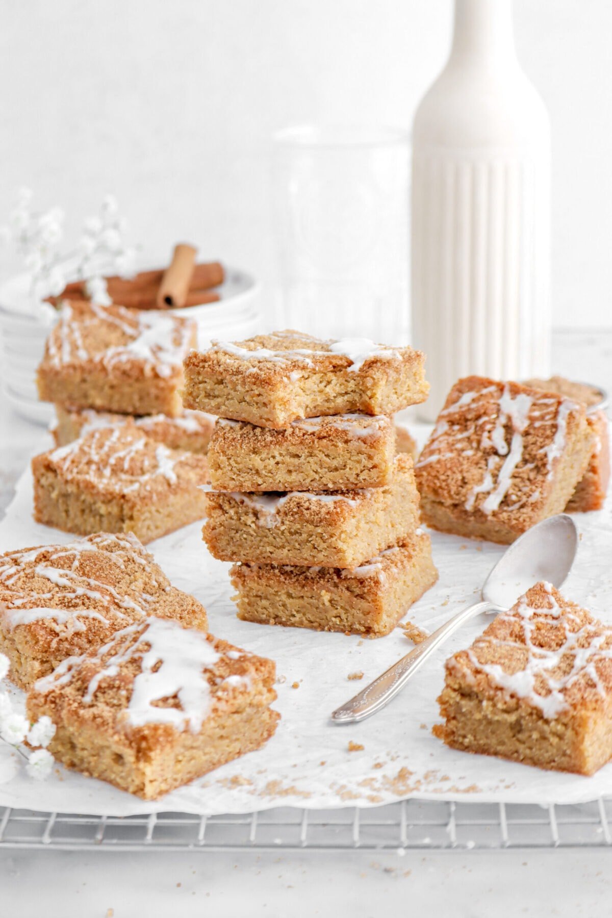 four stacked blondies with top blondie missing a bite on parchment lined cooling rack with more blondies around, a vase, and stack of plates behind.