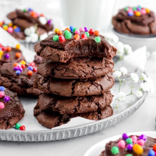 stacked cosmic brownie cookies on metal plate with more cookies around, white flowers, and a glass of milk behind.