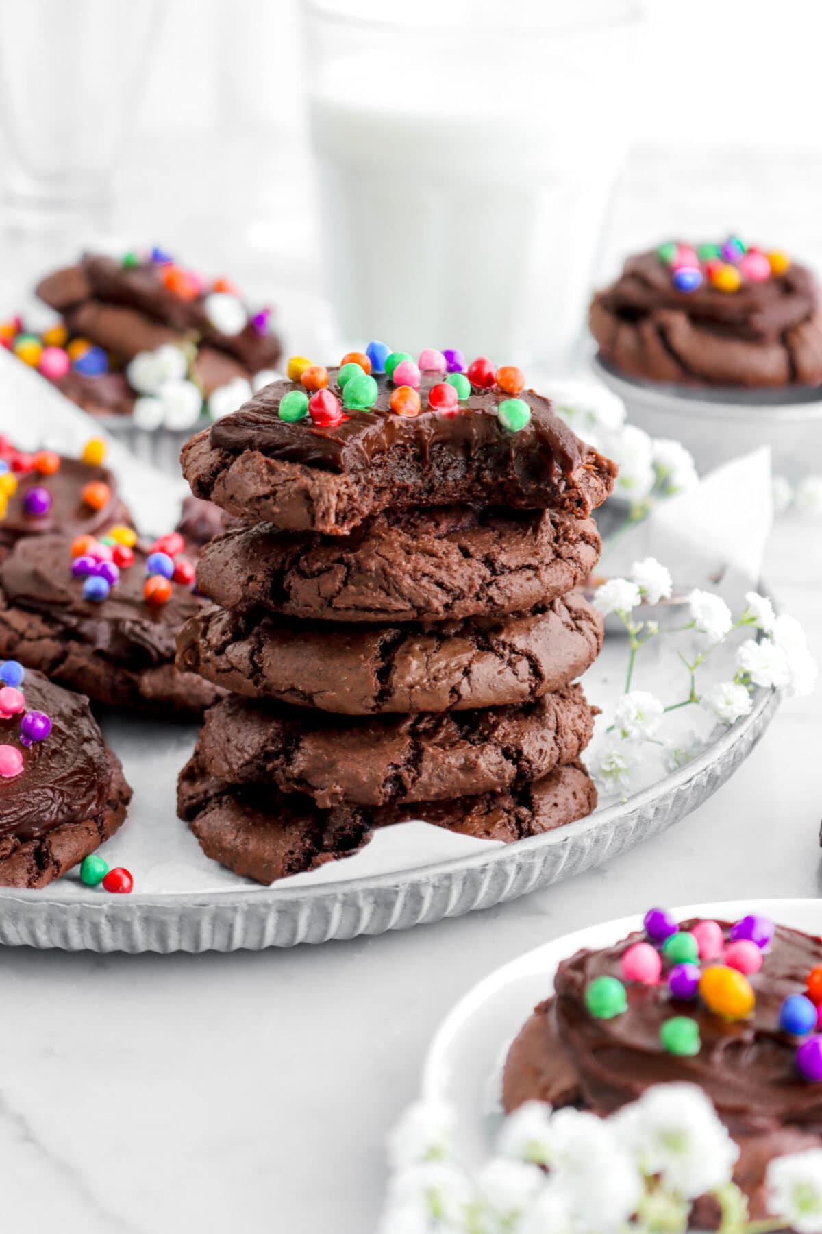 stacked cosmic brownie cookies on metal plate with more cookies around, white flowers, and a glass of milk behind.