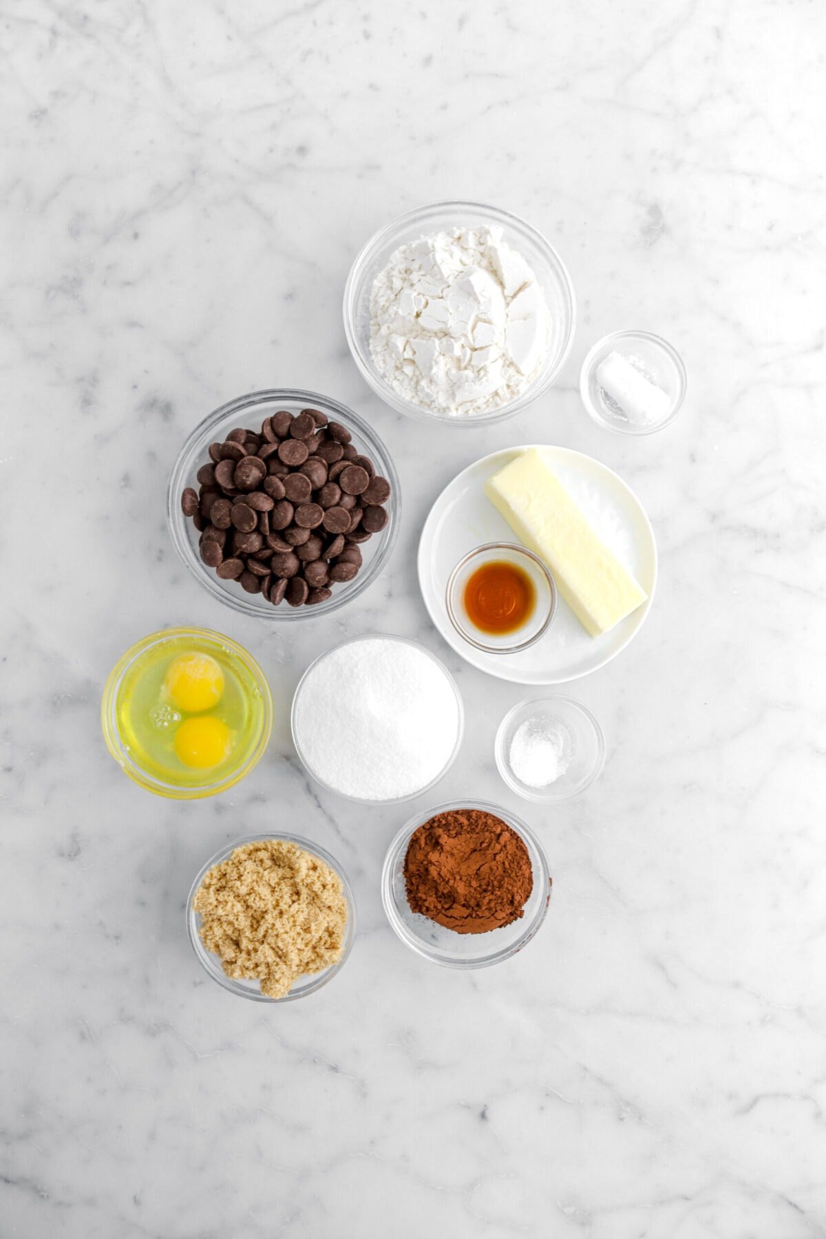 flour, baking powder, chocolate chips, butter, vanilla, sugar, salt, eggs, brown sugar, and cocoa powder on marble surface.