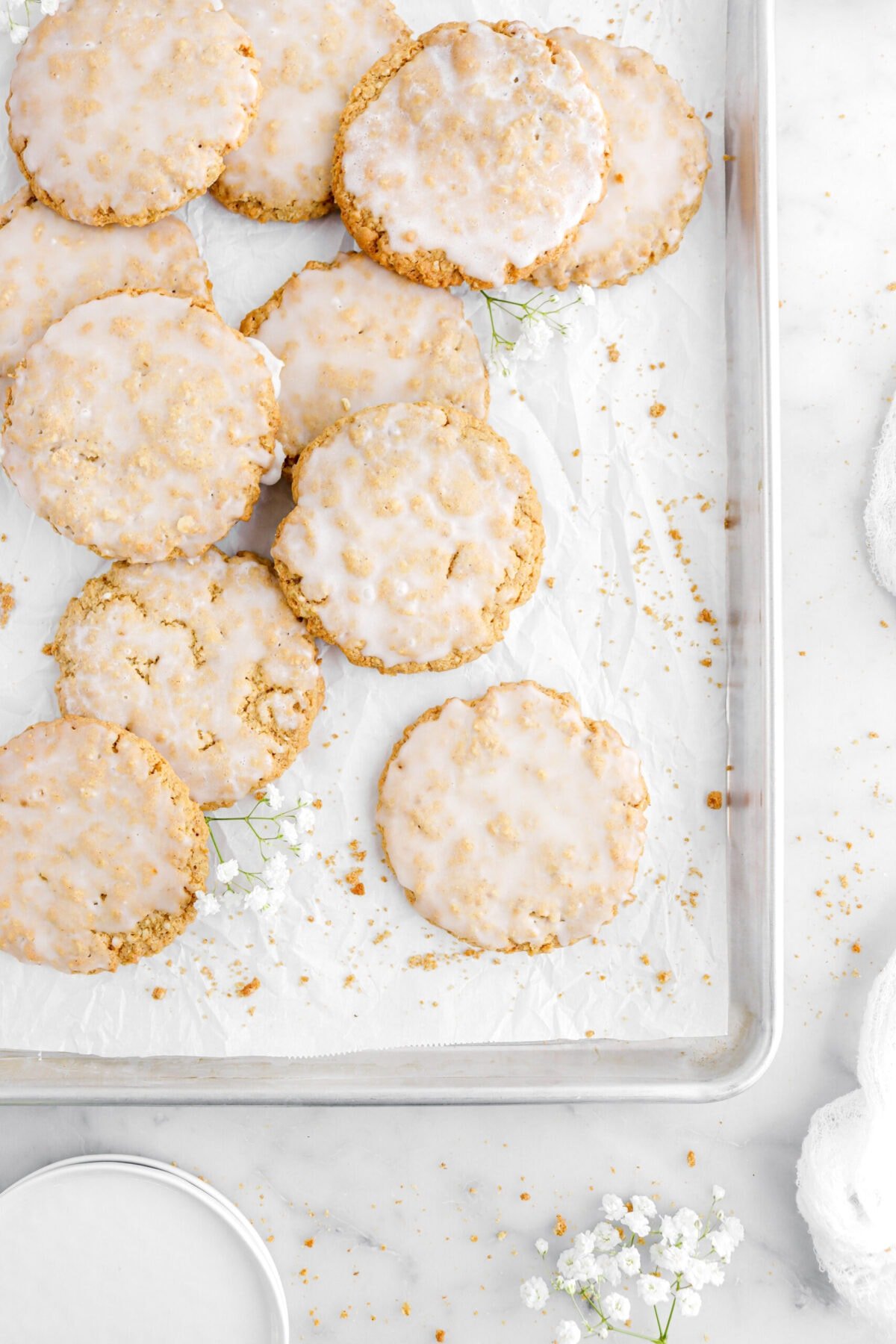 iced oatmeal cookies on lined sheet pan with cookie crumbs and white flowers around, a stack of white plates and white cheesecloth on marble surface next to pan.