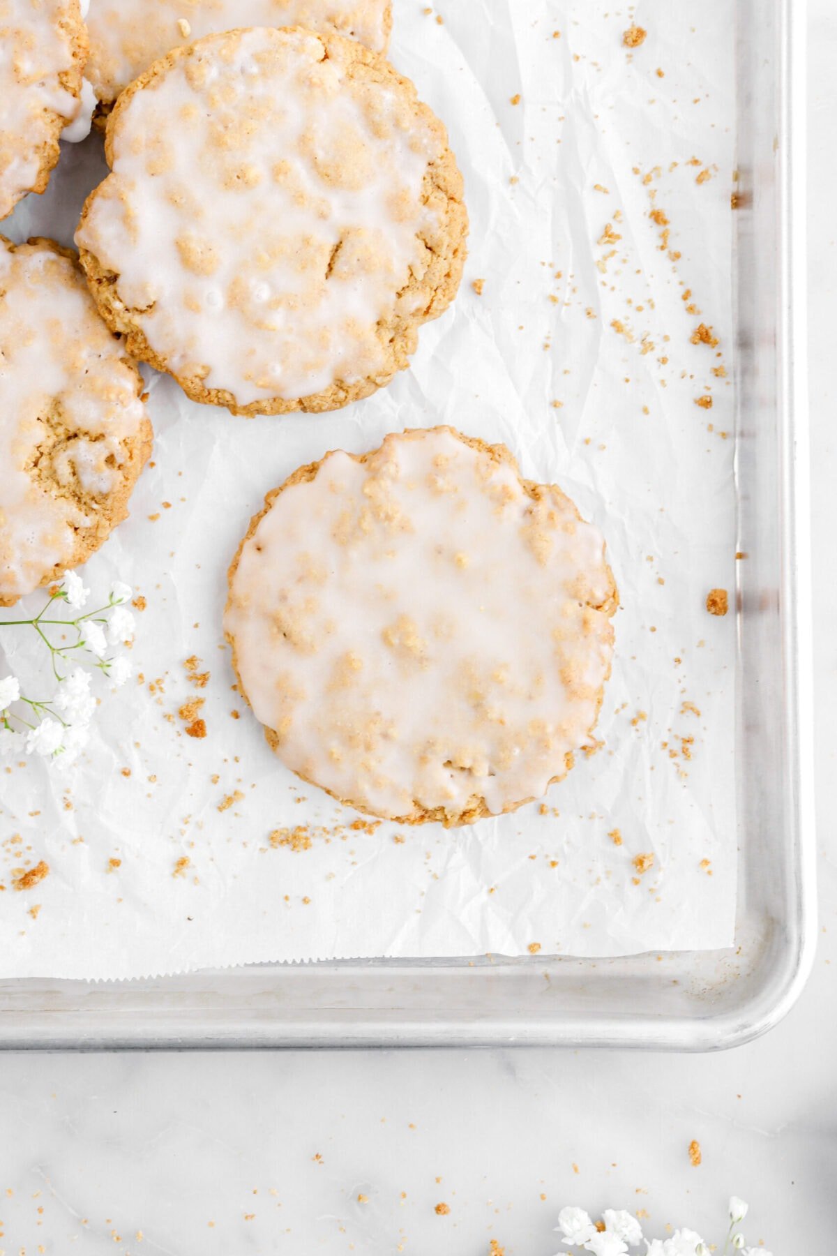 iced oatmeal cookie on lined sheet pan with more cookies beside and white flowers.