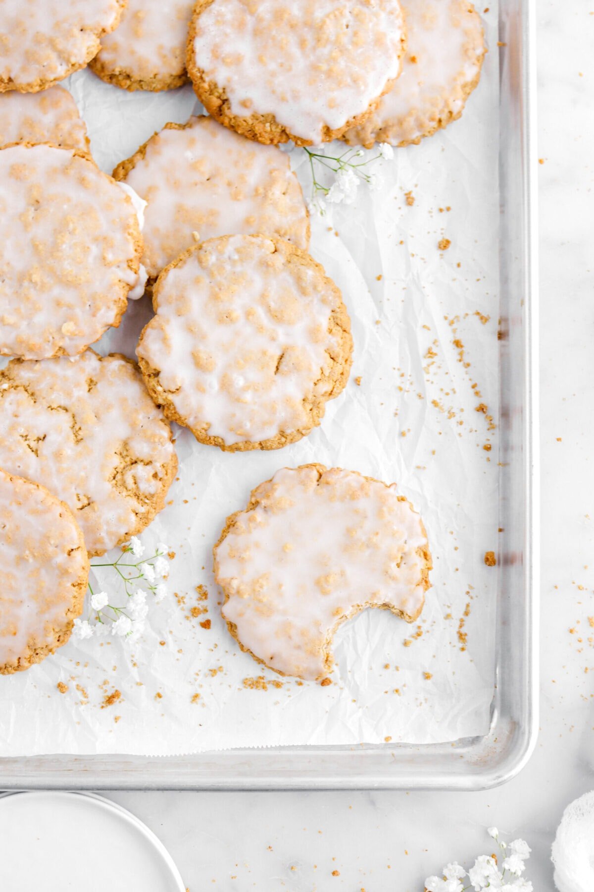 overhead shot of iced oatmeal cookies with bite missing on parchment lined sheet pan with cookie crumbs and white flowers.