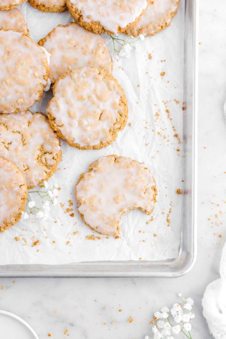iced oatmeal cookies on lined sheet pan with bite missing from one cookie with white flowers and cookie crumbs around.