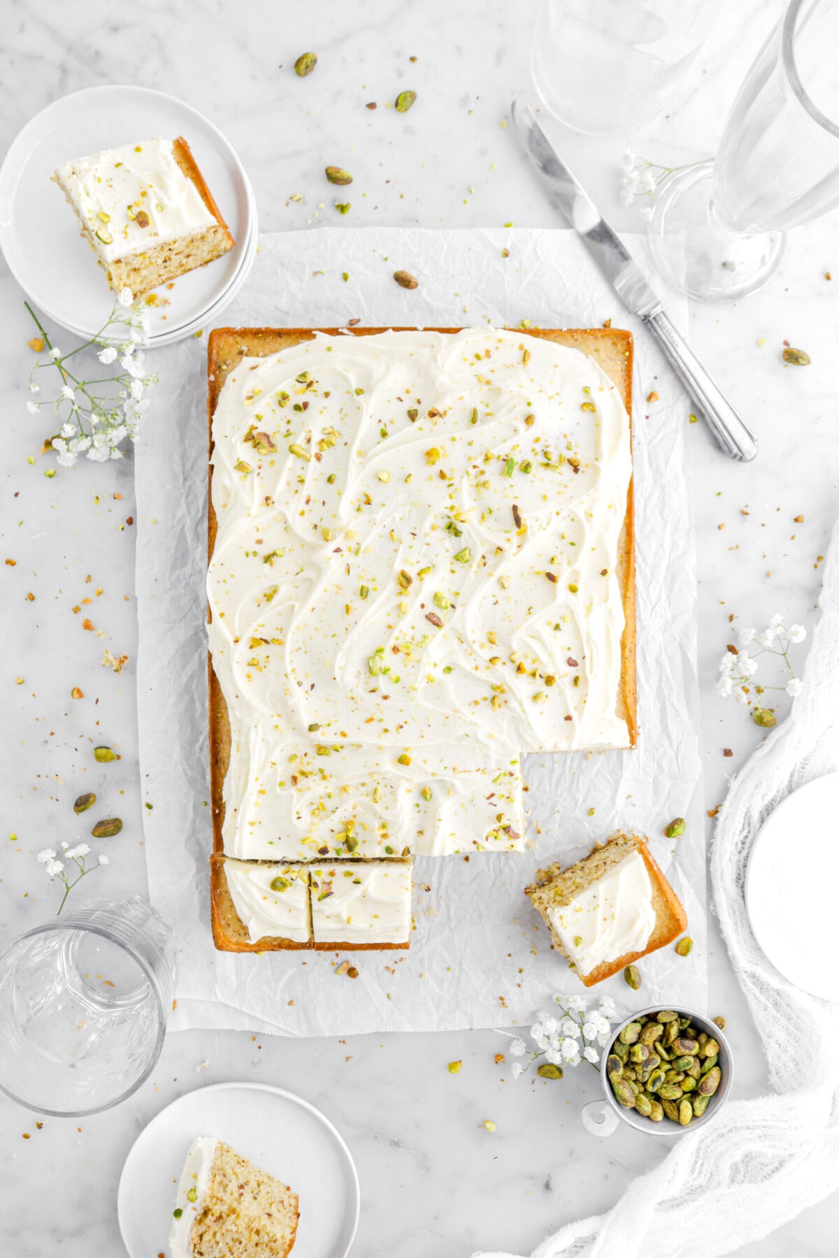 overhead shot of pistachio cake with slices cut into it with two slices on plates around cake with pistachios and white flowers around.