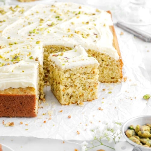 square slice of pistachio sheet cake in front of cake with another slice behind on parchment paper with pistachio crumbs and white flowers around.