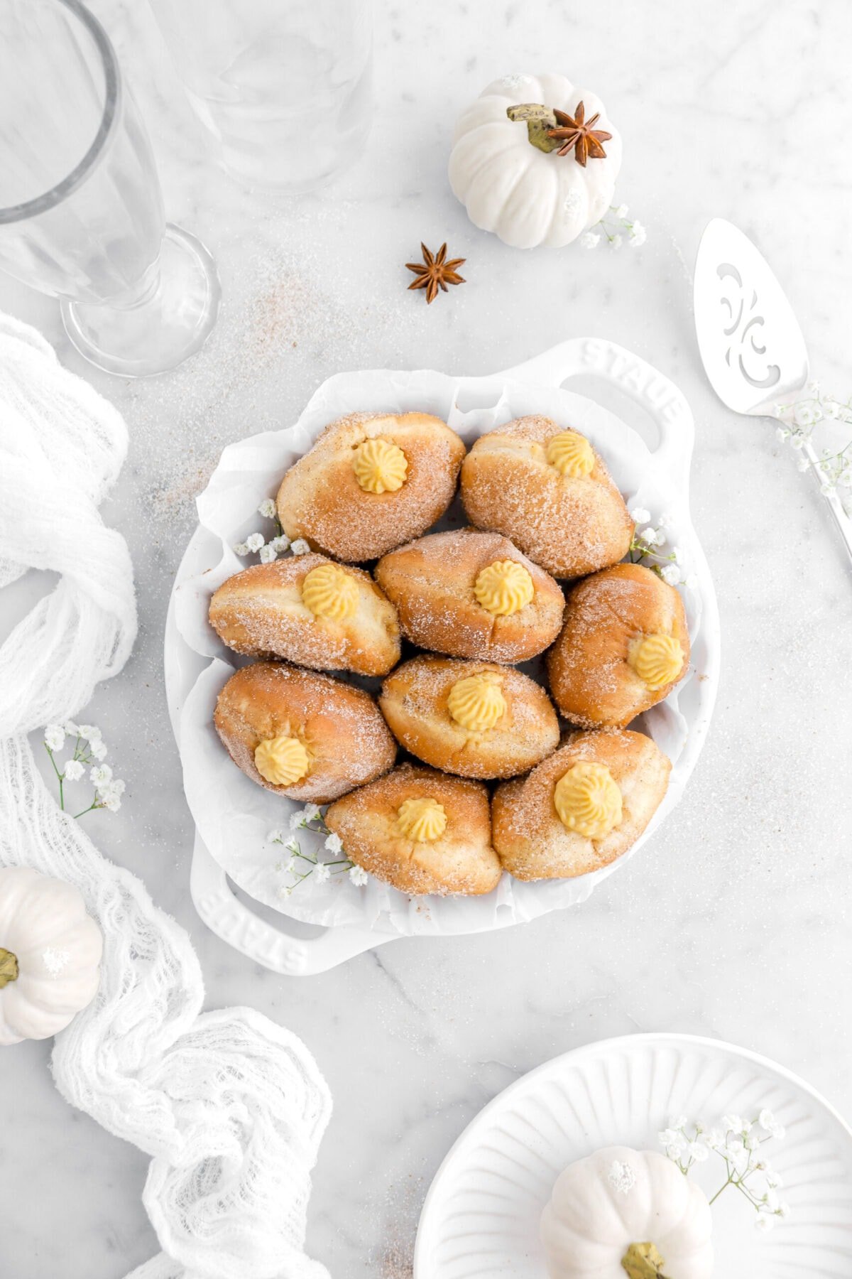 nine filled doughnuts in lined pie pan with white pumpkins, a white cheesecloth, and cake knife on marble surface.