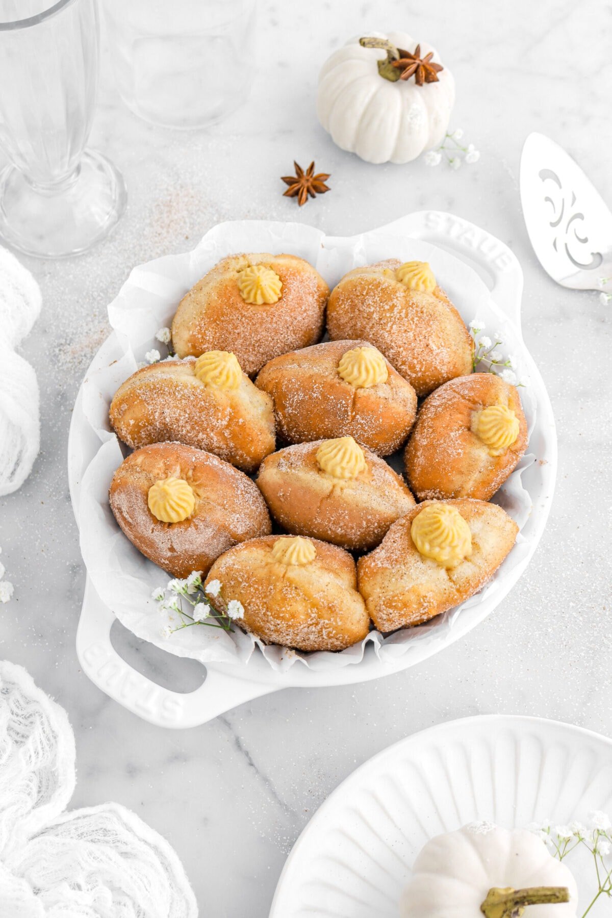 angled shot of nine pumpkin filled doughnuts in lined pie plate with flowers and white pumpkins around on marble surface.