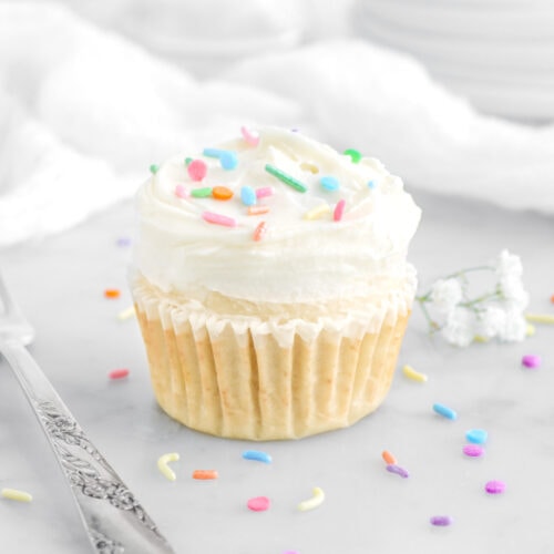 angled close up of vanilla cupcake on marble surface with sprinkles around, a fork beside, and white flowers behind.