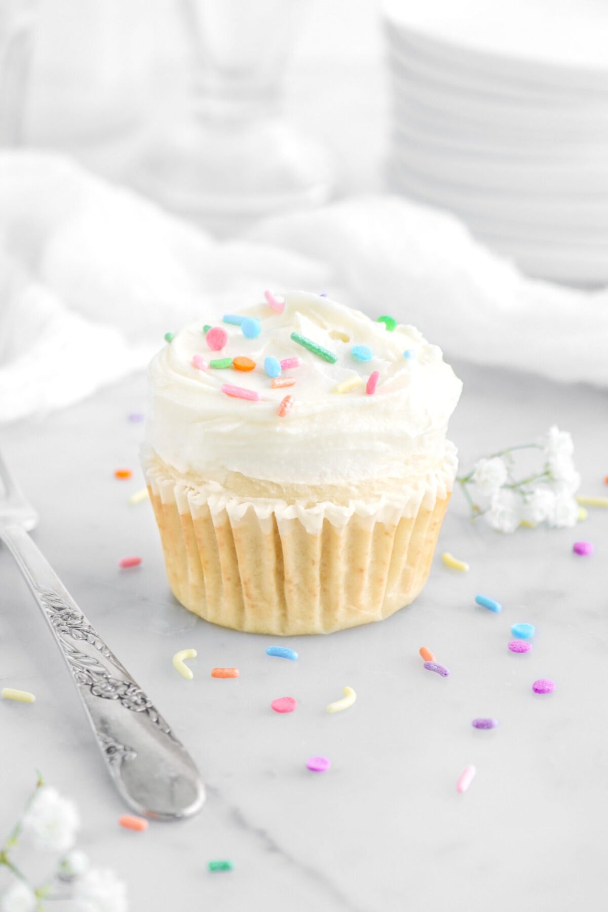 angled close up of vanilla cupcake on marble surface with sprinkles around, a fork beside, and white flowers behind.