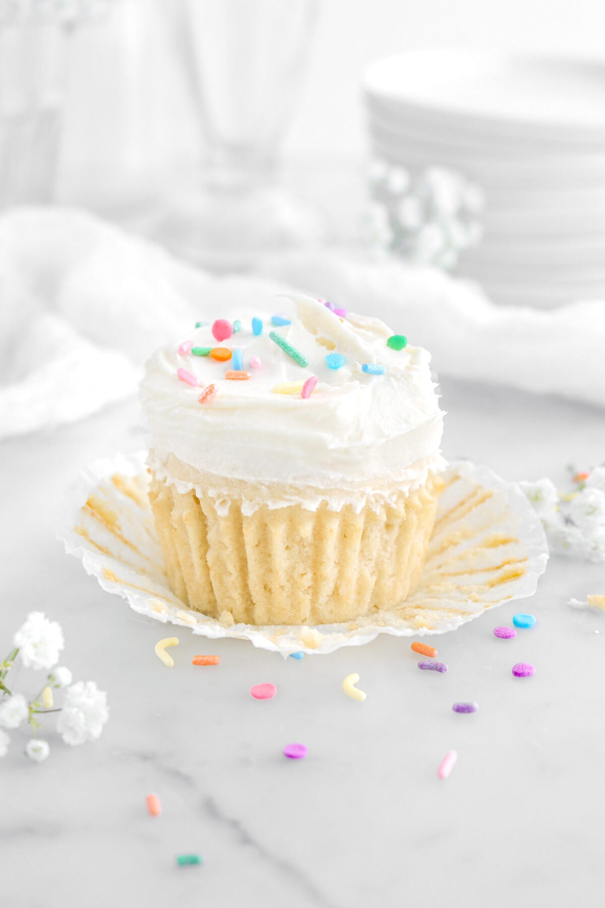vanilla cupcake with paper peeled away on marble surface with rainbow sprinkles and white flowers around.