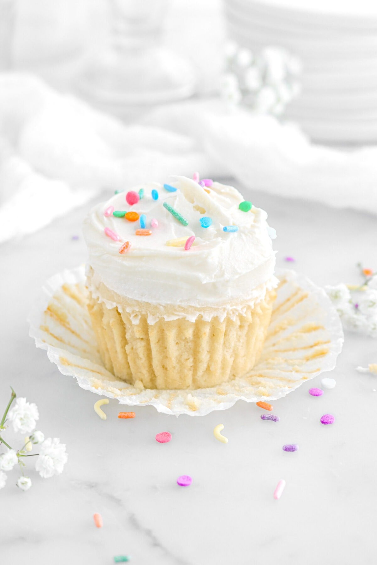 angled close up of vanilla cupcake with paper peeled away with flowers and sprinkles around on marble surface.