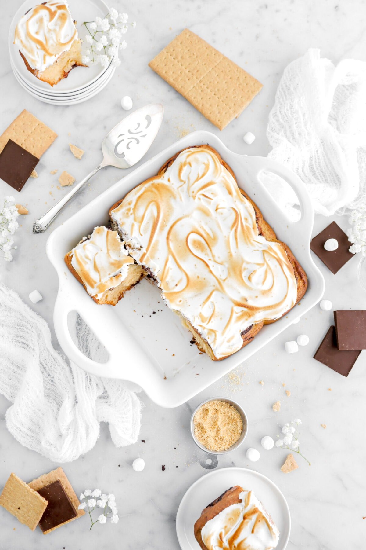 two s'mores cinnamon rolls on white plates beside casserole with seven rolls in white casserole on top of a white cheesecloth.