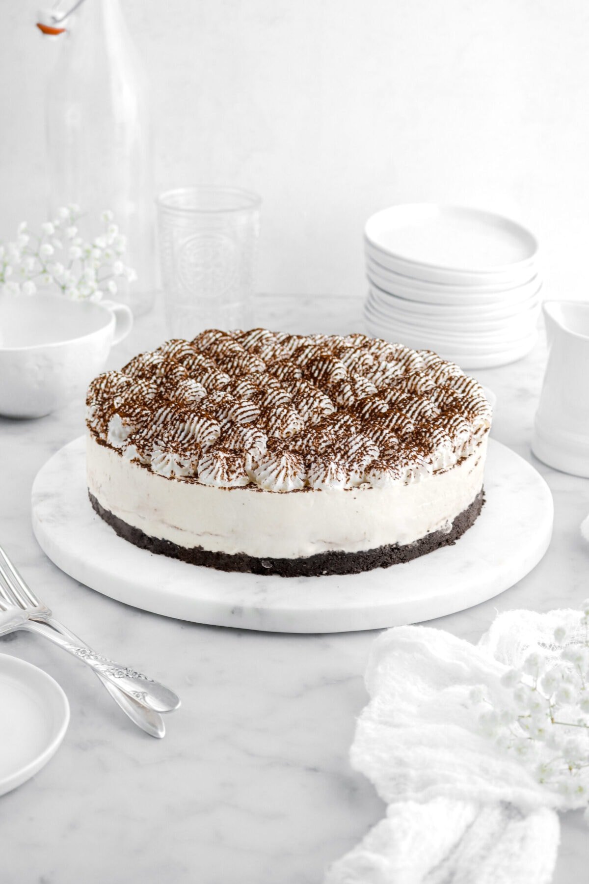 tiramisu cheesecake on marble serving tray with white flowers, two fork beside, and a stack of white plates behind.