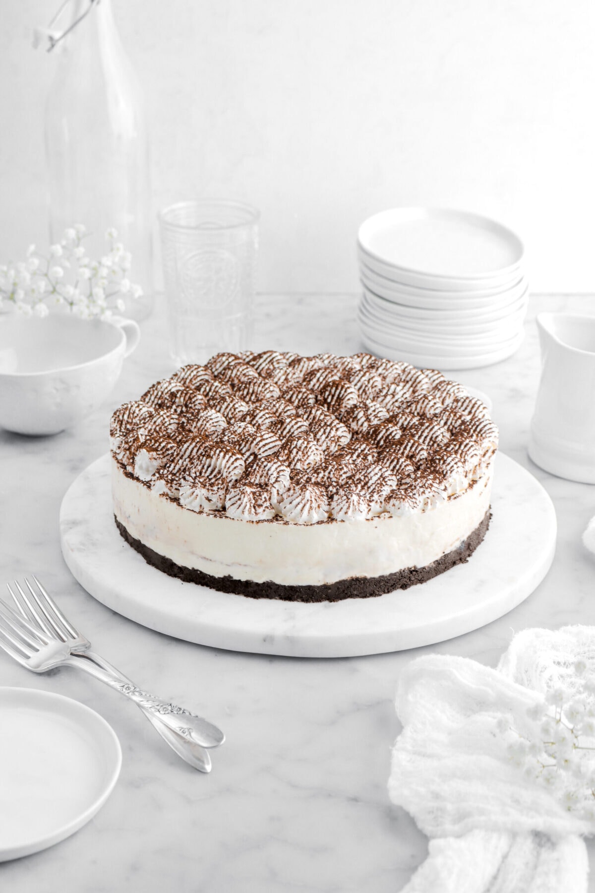 angled shot of tiramisu cheesecake on round marble tray with two forks beside, a white cheesecloth, white flowers, a stack of plates, and two empty glasses behind on marble surface.
