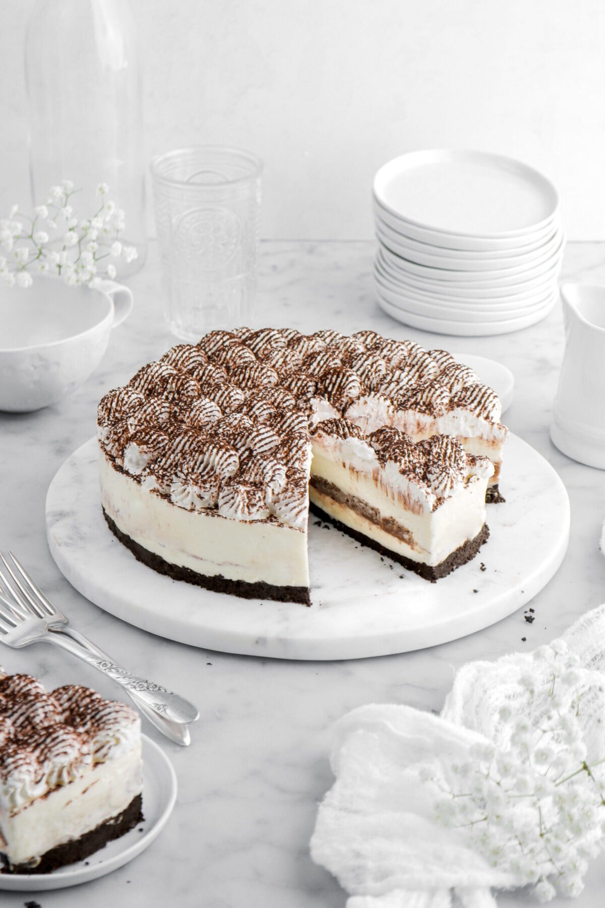tiramisu cheesecake with slice cut into it on round marble tray with another slice on small white plate beside and stack of plates behind.