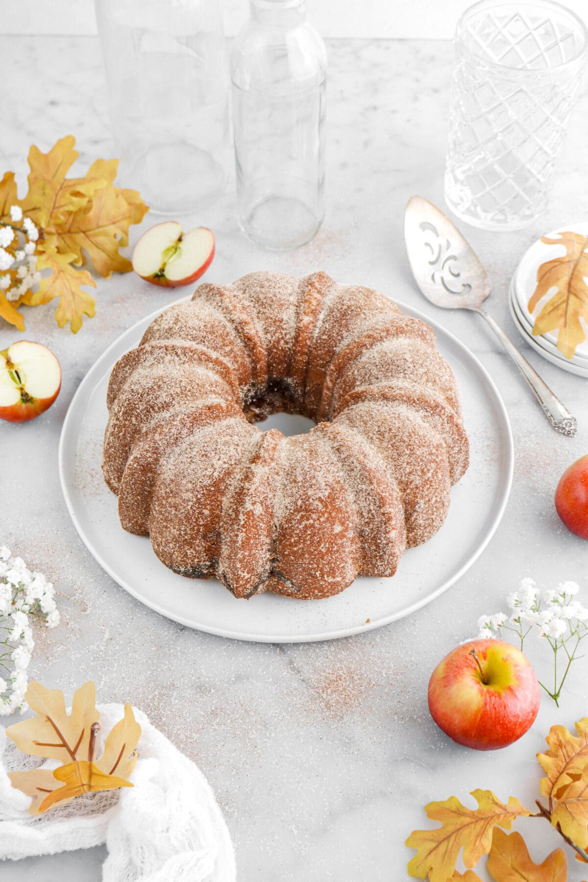 angled image of apple cider donut cake on white plate with yellow leaves and apples around on marble surface.