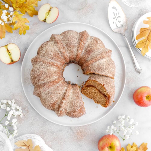 overhead image of cut apple cider donut cake with slice on white plate beside with yellow leaves, flowers, apples, a cake knife, and empty glasses around.