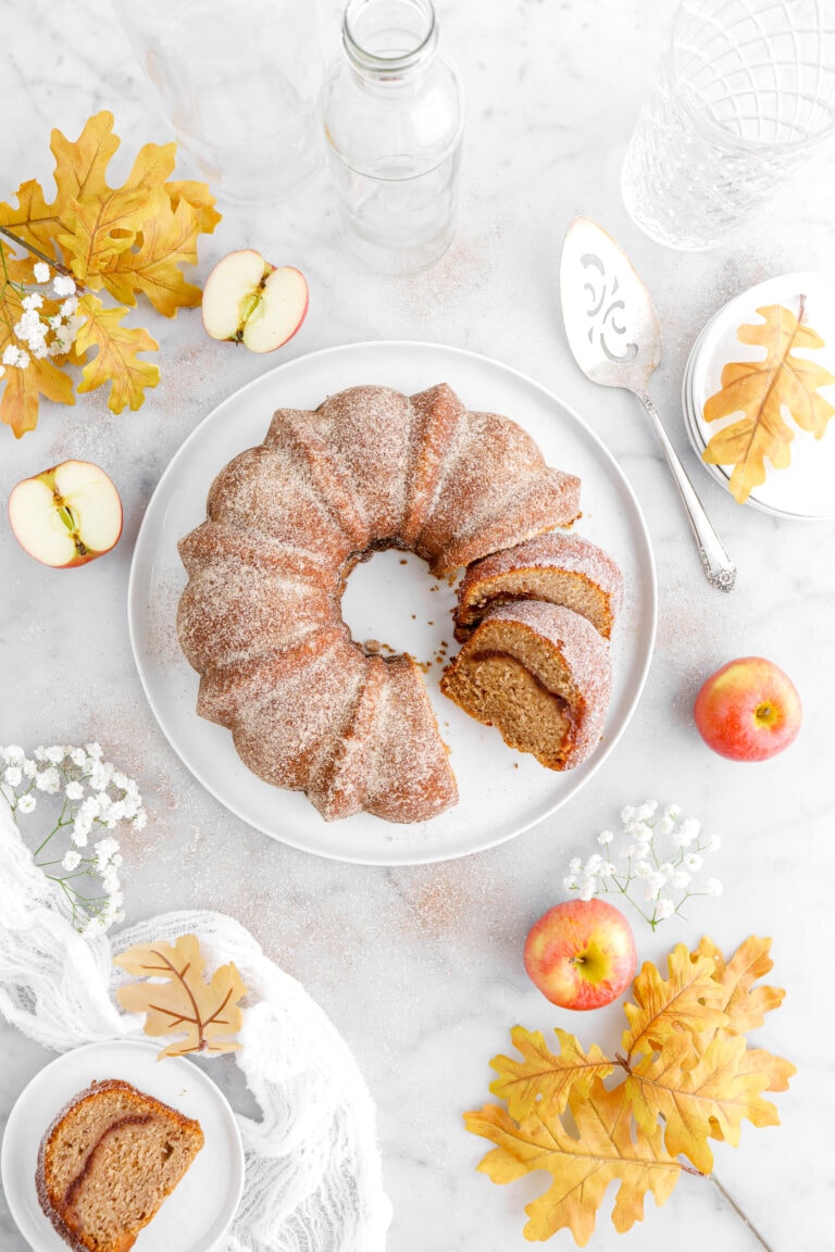 overhead image of apple cider donut cake with two slices cut into cake on white plate with third slice on smaller white plate beside on marble surface with apples, white flowers, and yellow leaves around.