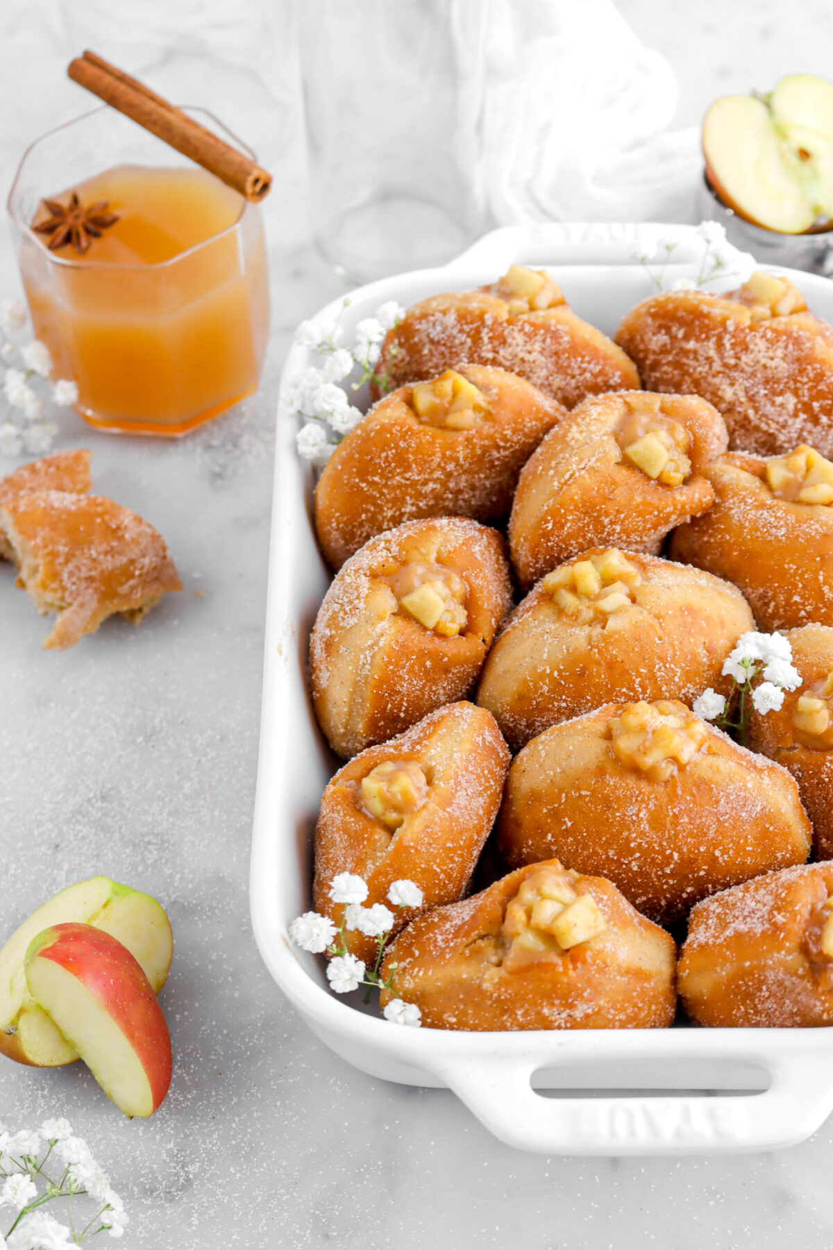 cropped image of apple pie filled doughnuts in white casserole on marble surface with apple slices and a glass of cider beside.