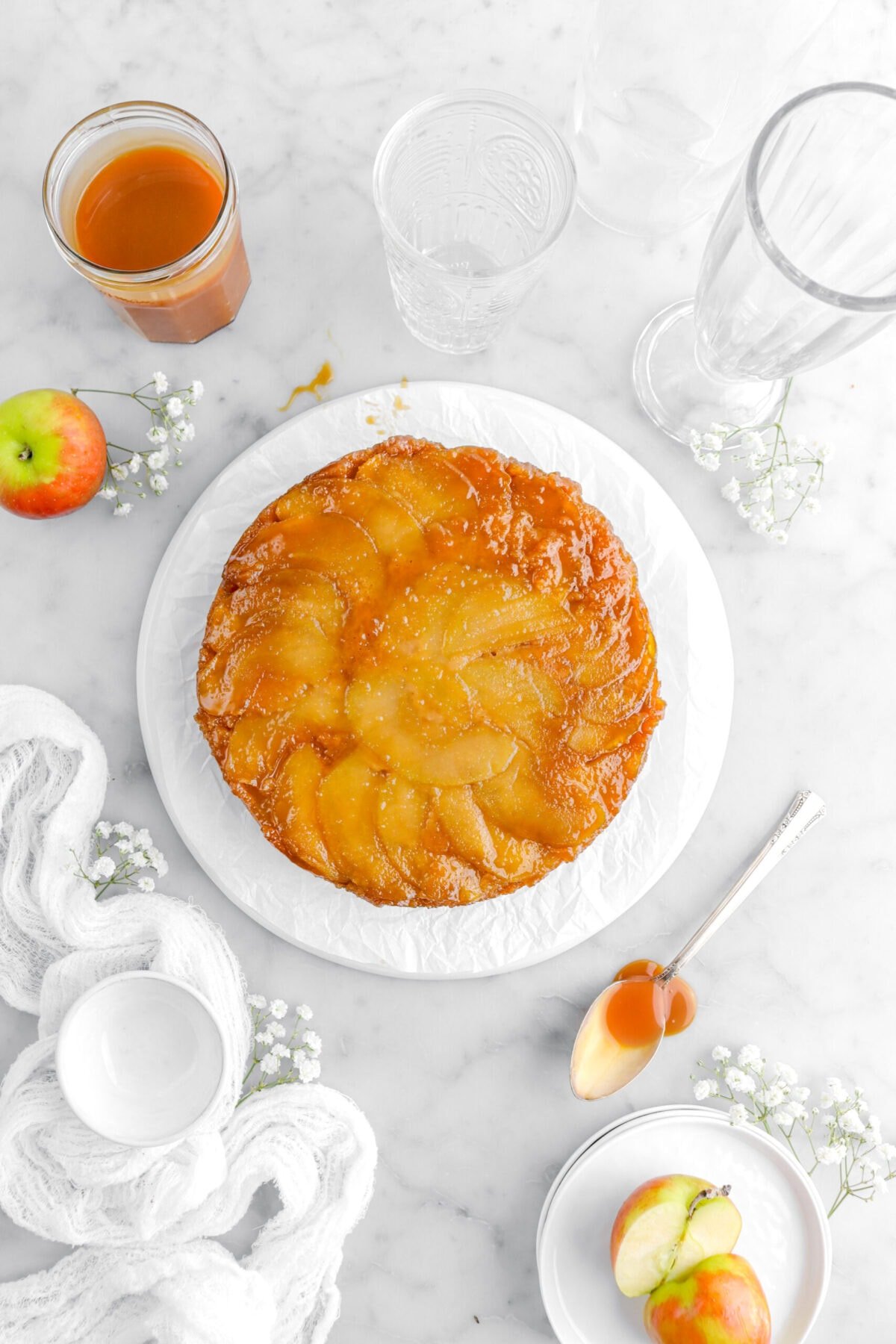 caramel apple upside down cake on parchment paper with spoon and jar of caramel beside, apples, flowers, and empty glasses around on marble surface.