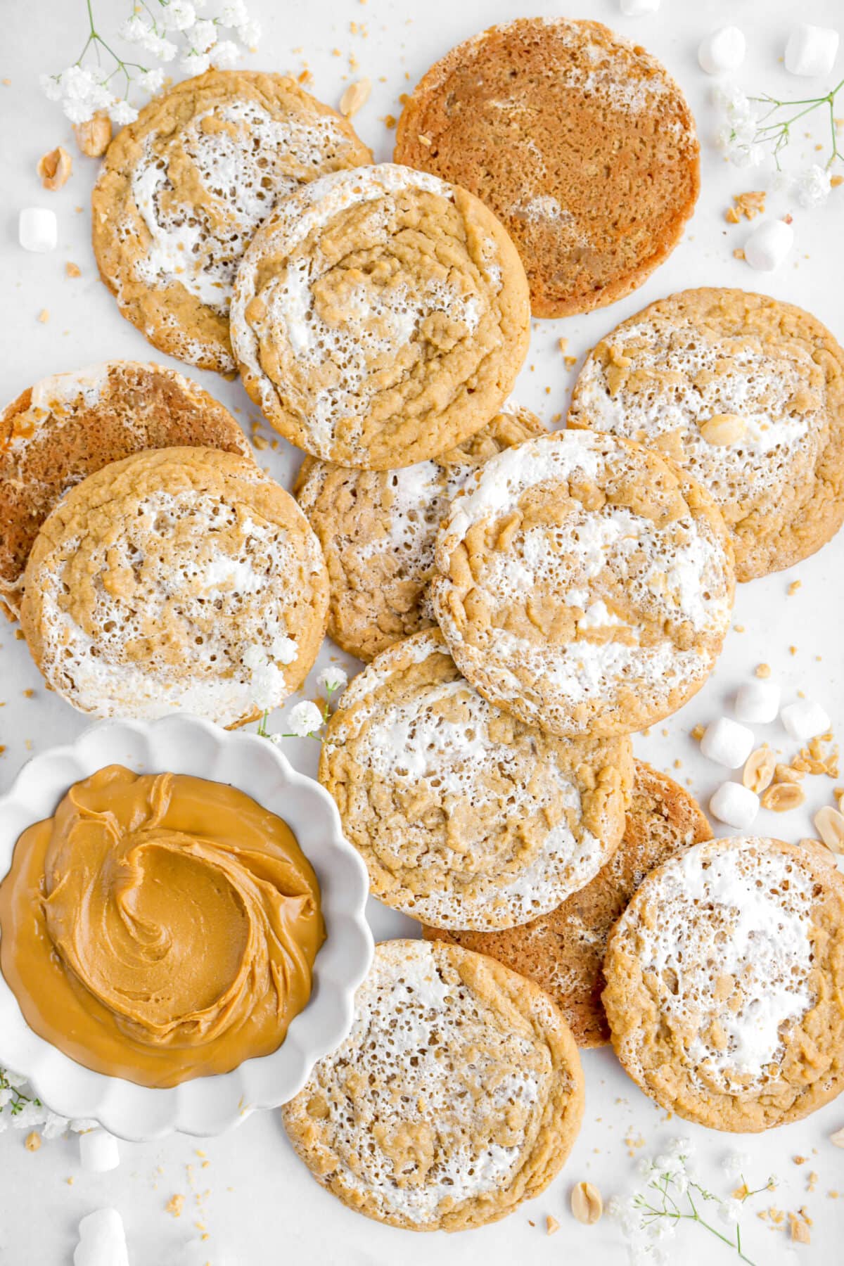 peanut butter marshmallow cookies piled on parchment paper with bowl of peanut butter beside and mini marshmallows, peanuts, and white flowers around.