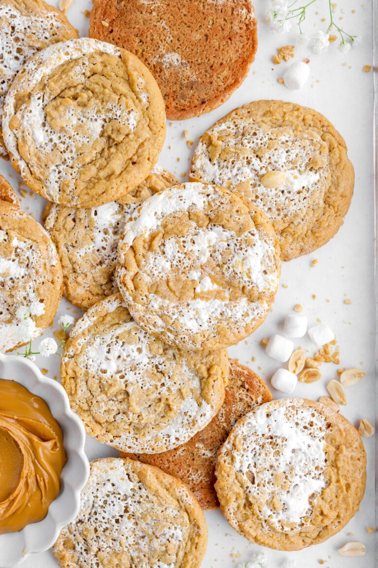 overhead image of peanut butter marshmallow cookies on parchment paper with peanuts, mini marshmallows, and white flowers around.