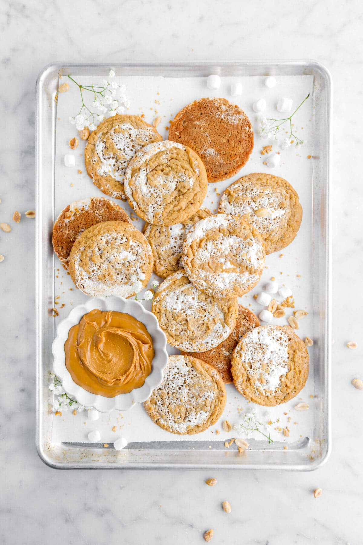 overhead image of fluffernutter cookies on lined sheet pan with small bowl of peanut butter with mini marshmallows, peanuts, and white flowers around.