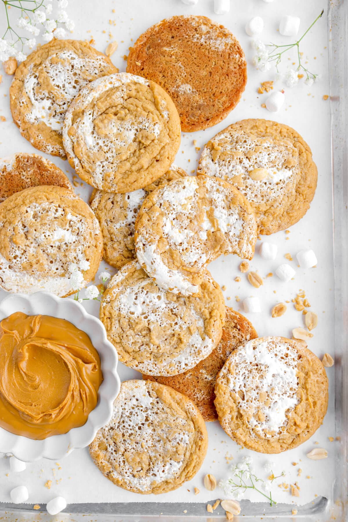 cropped image of peanut butter marshmallow cookies on lined sheet pan with bowl of peanut butter, mini marshmallows, and white flowers.