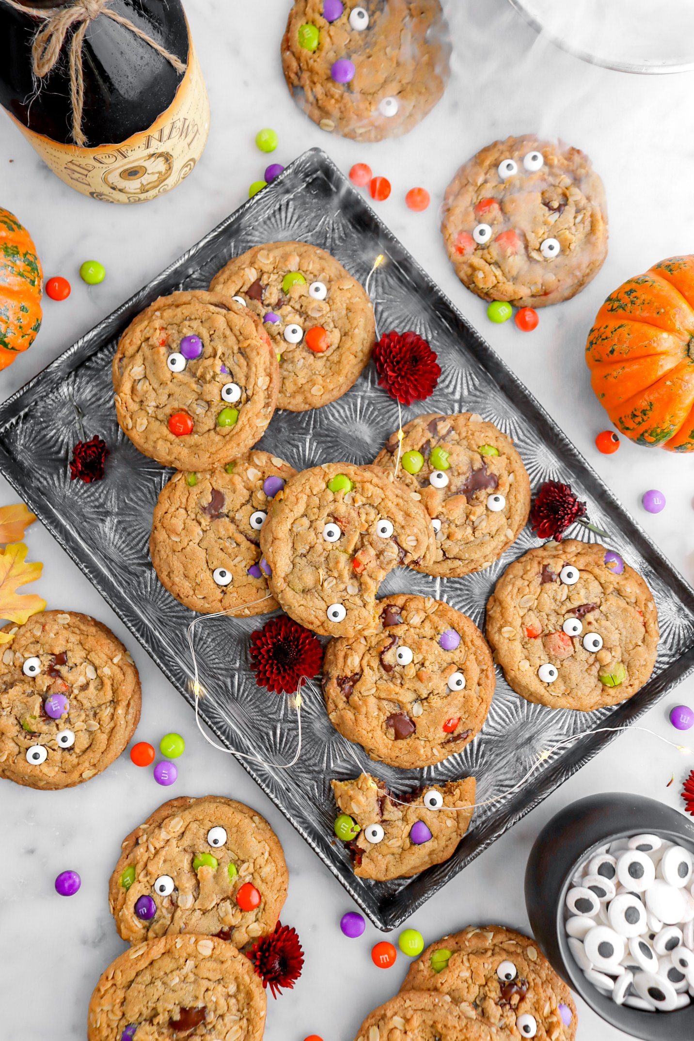 overhead image of monster cookies on dark sheet pan with one cookie missing a bite, a string of lights, red flowers, and more cookies around on marble surface.
