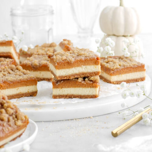 two staked pumpkin maple bars on marble surface with more bars around and a stack of white pumpkins behind and white flowers around.