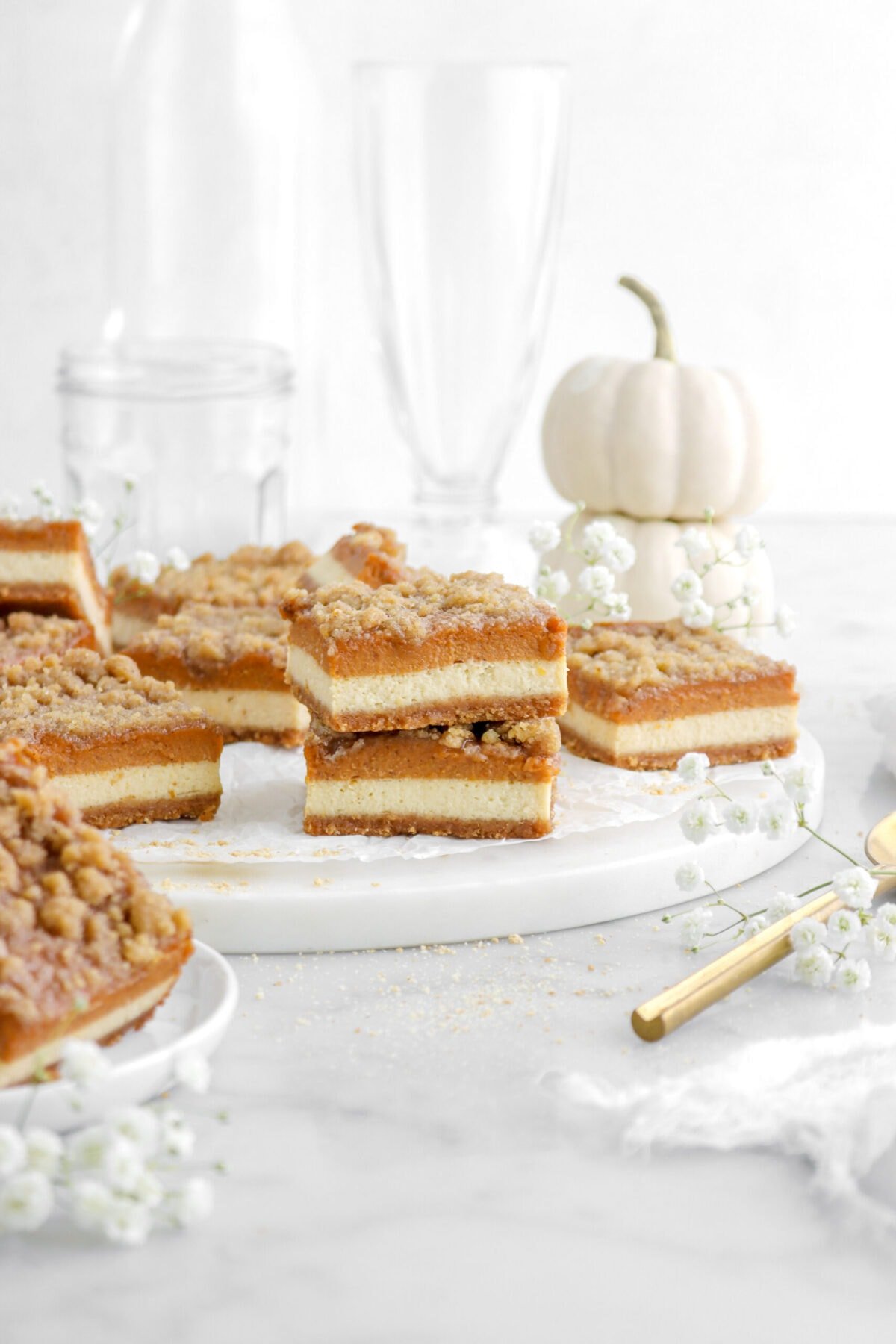 front shot of pumpkin cheesecake bars stacked on marble serving tray with more pumpkin bars behind and white flowers around, empty glasses, and a stack of white pumpkins behind.
