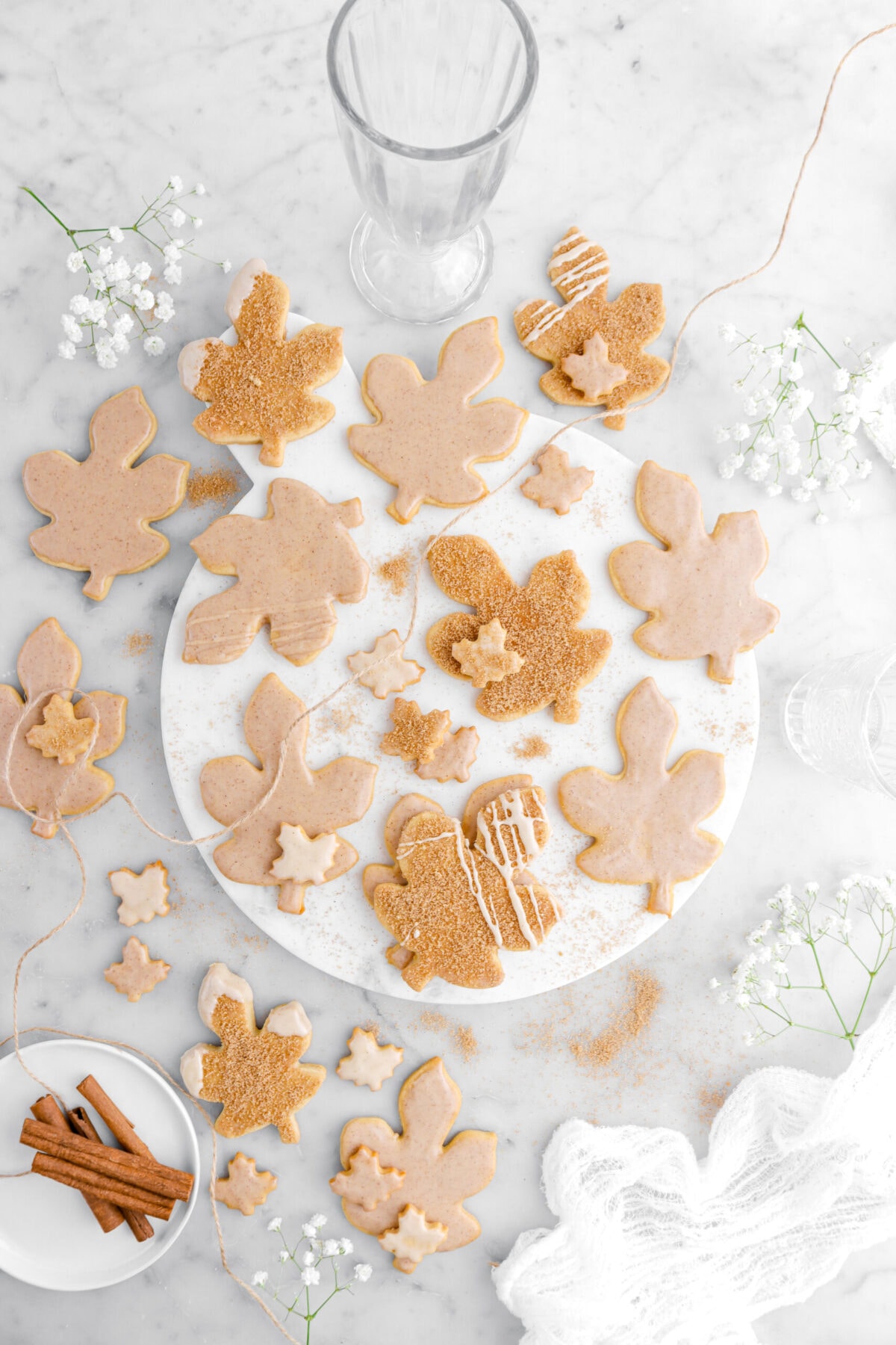 overhead image of maple cookies on marble surface with cooking twine, cinnamon sticks, white flowers, cheesecloth, an empty glass on marble surface.
