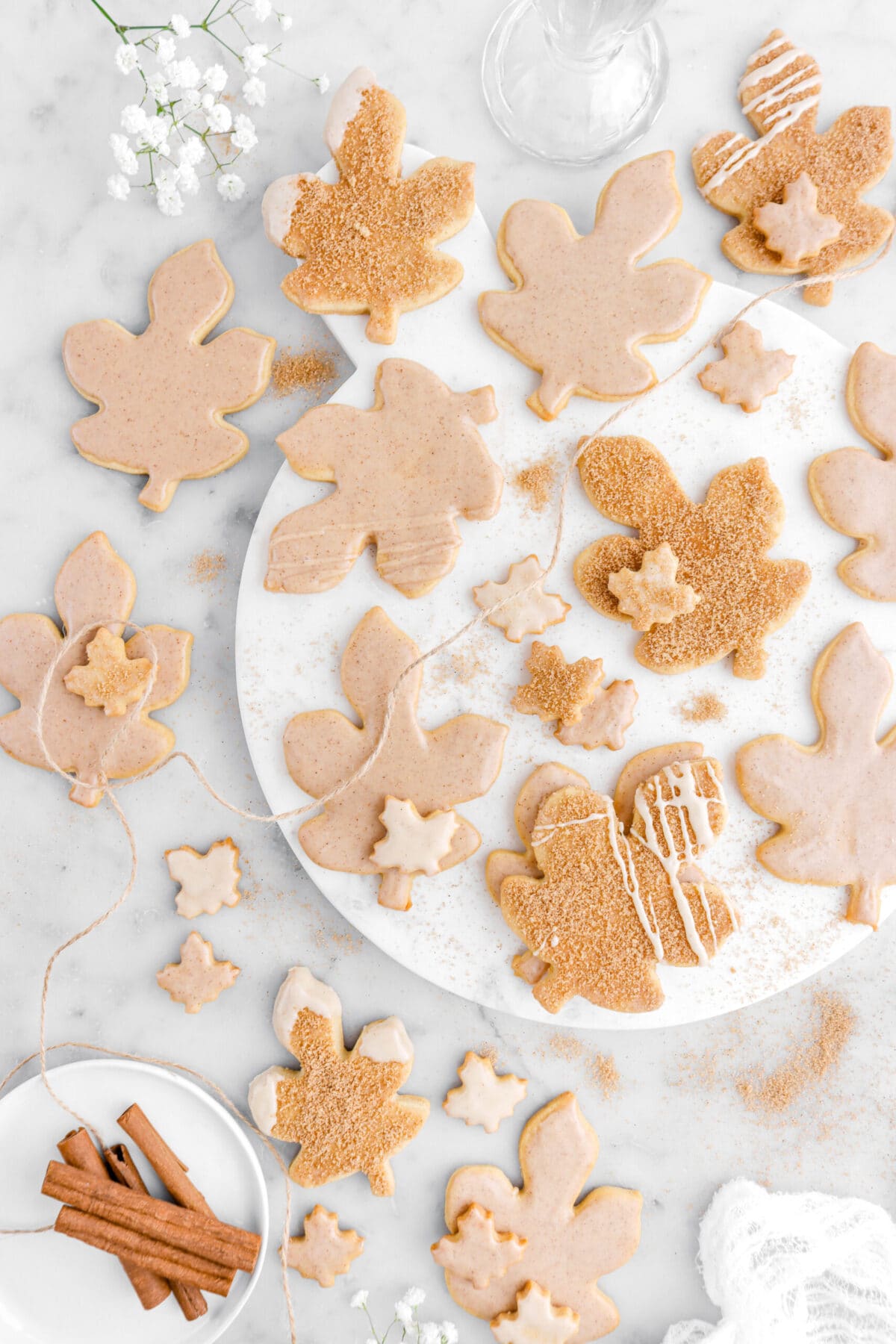 cropped overhead image of maple leaf shaped cookies on marble surface with cinnamon sugar around, cinnamon sticks, and cooking twine on marble surface.