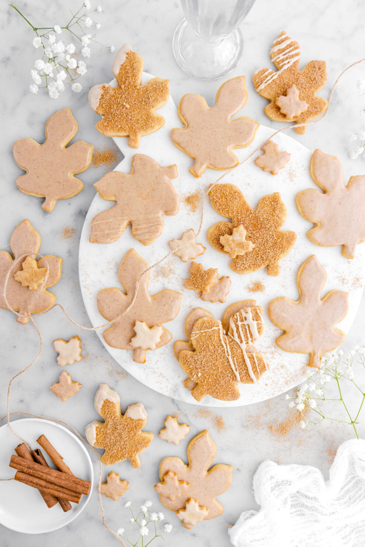 cropped image of iced maple leaf cookies on marble tray with white flowers, cinnamon sugar, and cinnamon sticks around.