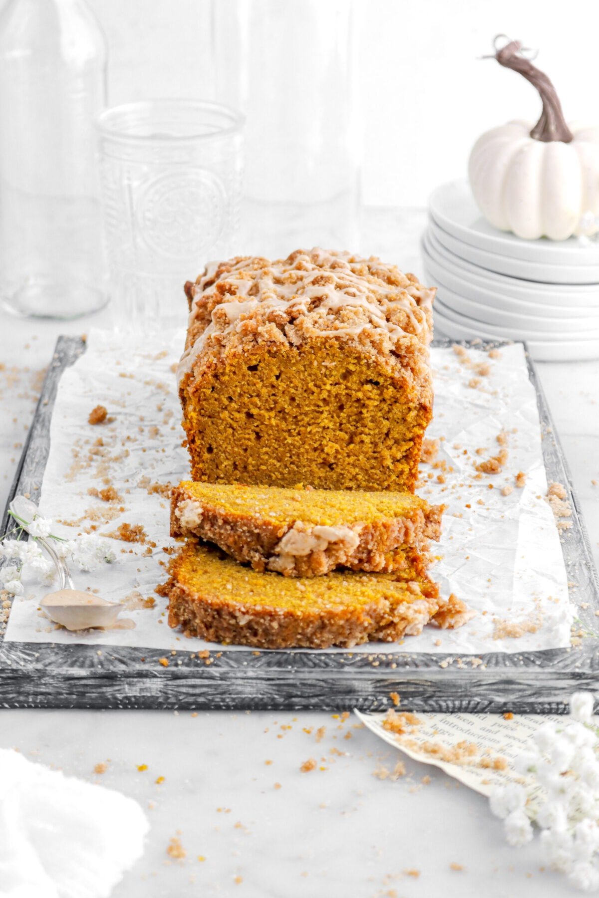 pumpkin loaf on upside down sheet pan with two slices laying in front with empty glasses and stack of plates behind with white flowers and streusel crumbs around.