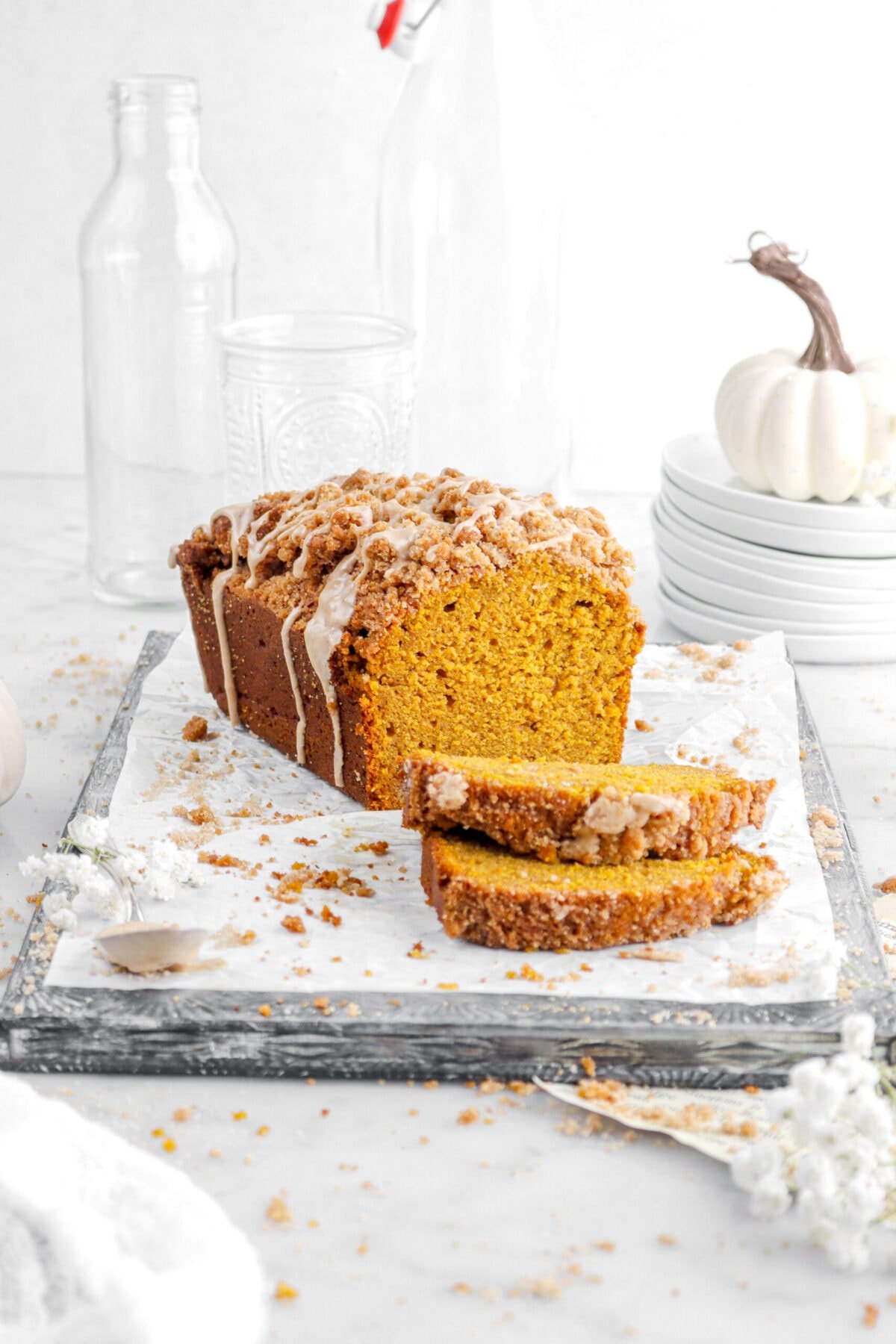 angled pumpkin loaf on upside down sheet pan with two slices of bread laying in front with stack of plates, small pumpkin, and empty glasses behind with streusel crumbs and white flowers around.