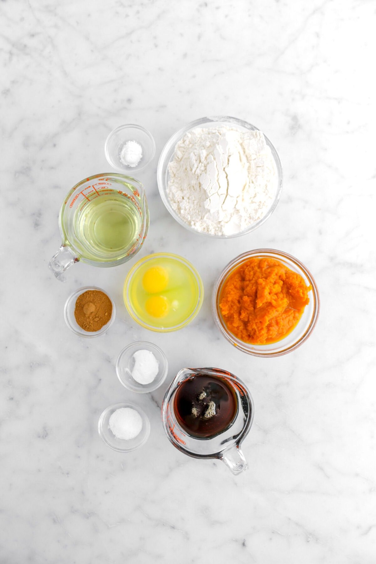 baking powder, flour, oil, eggs, spice, baking soda, salt, maple syrup, and pumpkin purée on marble surface.