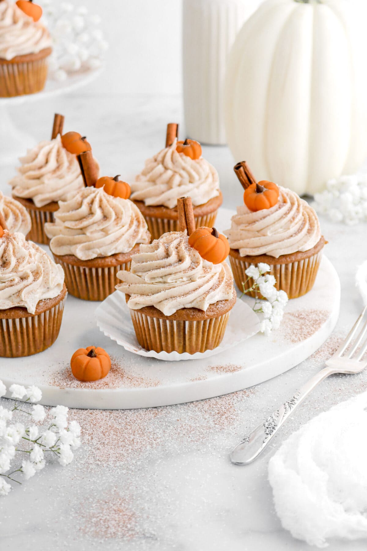 angled image of pumpkin cupcakes on marble board with fondant pumpkins, cinnamon sticks, and spiced sugar on top with white flowers and a fork beside.