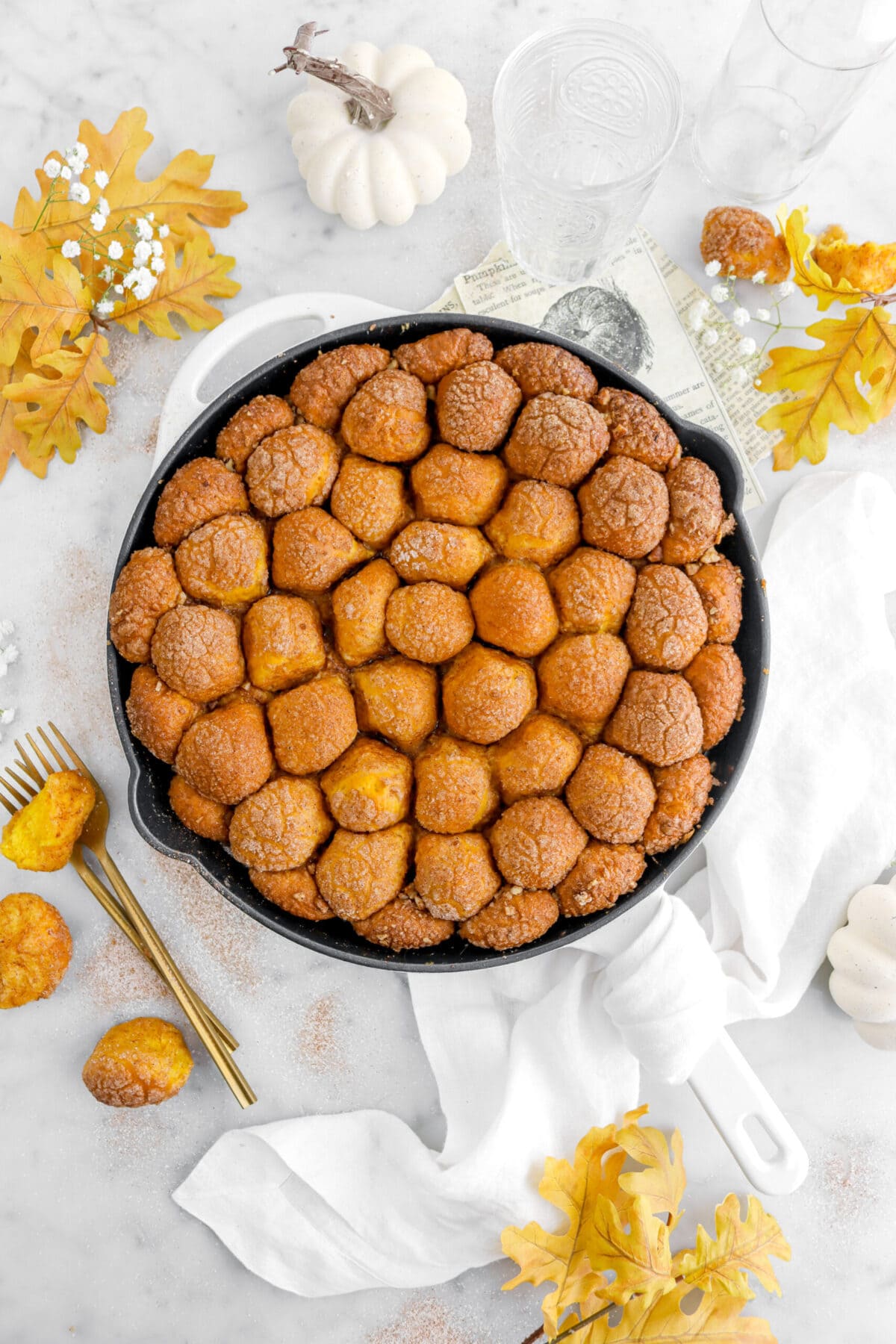skillet pumpkin monkey bread on marble surface with yellow leaves, white pumpkin, and white flowers around.