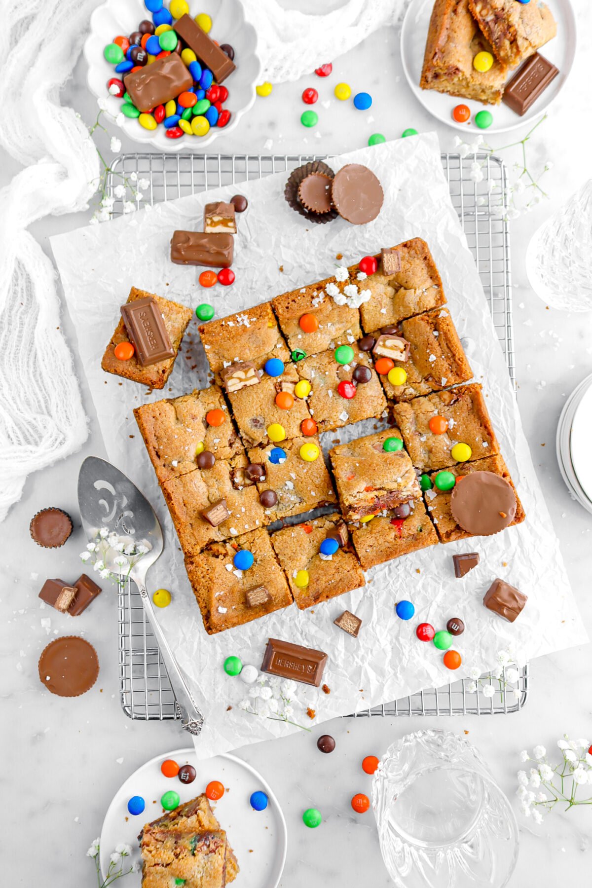 cookie bars on parchment paper on cooling rack with candies and white flowers around, and candy on top of cookie bars.
